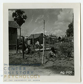 Three Indian refugees crouching in a dirt and grass area of Azadgarh refugee camp. Behind them is the camp itself and three buildings are visible. “AG. 500.” is written in white in the bottom right corner.