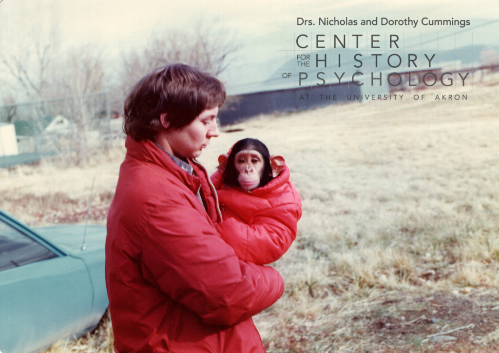 A photograph of a man with dark hair holding a young chimpanzee. They are both wearing red winter coats. There is a blue car in the background and mountains in the distance.