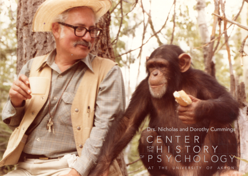 A photograph of a man with a moustache wearing a tan hat, tan vest, and plaid shirt is sitting next to a chimpanzee. The man is holding a cup and the chimpanzee is holding bread. They are sitting outdoors.