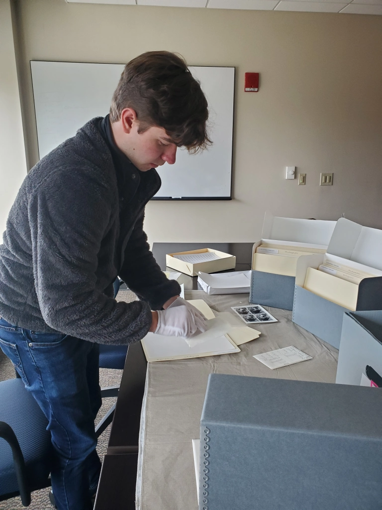 A photograph of a male student assistant with short dark hair in front of a table. The table holds boxes and folders. The student is wearing white gloves and handling photographs.