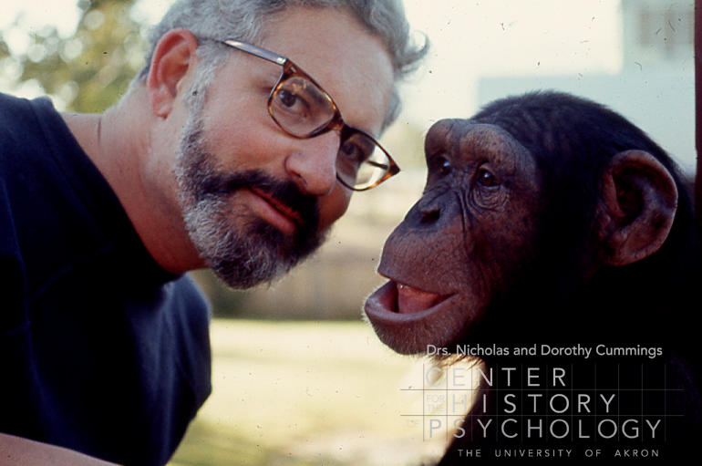 A photograph of a man with a beard and glasses and navy blue shirt with his head close to a chimpanzee. It is a head-and-shoulders view of both the man and chimpanzee.