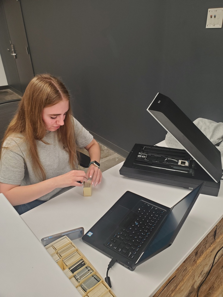 A photograph of a female student with long dark blonde hair is sitting at a desk in front of a computer and scanner. She is handling photographic slides in a small yellow box.