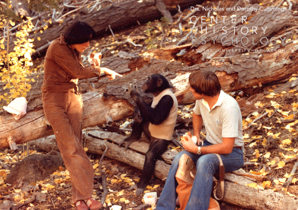 A digitized photograph of two people and a chimpanzee sitting on large logs outside in the woods. The woman on the left is holding a sandwich and pointing toward the chimp.  The chimp is wearing a white sweater vest and using sign language toward the woman.  The man is holding a sandwich and looking toward the chimp.