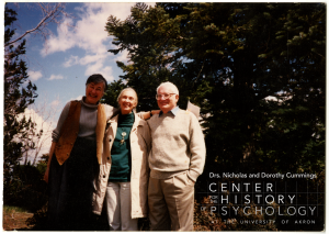 A photograph of three people facing the camera with their arms around each other. Beatrix T. Gardner is on the left, Jane Goodall is in the middle, and R. Allen Gardner is on the right. They are outdoors in front of a large pine tree with blue sky in the background.